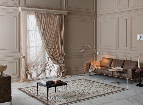 This picture shows a modern, luxurious bedroom with terracotta-colored walls. The floor is covered in carpet and there are upholstered chairs and a chaise lounge in the corner. The bed is large and has a white leather headboard. A sleek black dresser is against the wall on one side, and a mirror hangs on the opposite wall. There are windows with sheer curtains providing natural light, and several potted plants for added warmth.