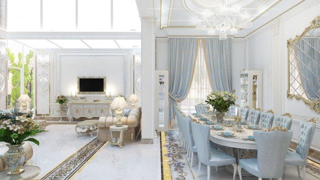 This picture shows a luxurious, opulent living room design. The room has richly colored walls with intricate gold and beige accents. There are two large, ivory-colored chaise lounge chairs in the center of the room, facing a white marble fireplace flanked by two mirrors. A round, ornate crystal chandelier hangs from the ceiling. The floor is covered in a light beige and gold carpet and the walls have multiple pieces of art decorating them.