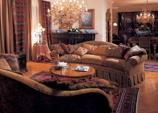This picture shows a luxurious bedroom with a stunning marble fireplace. The room is decorated in a neutral palette, with a taupe colored sofa and plush ottoman at the center of the room. There are several ornate gold accents mixed with white and cream furniture, intricate décor pieces, and lush carpets on the floor. The walls are adorned with beautiful paintings and a large mirror hangs above the fireplace, creating an elegant and opulent feel.