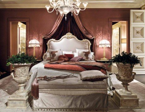 This picture shows a luxurious bedroom decorated in modern style with neutral colors. The room has a contemporary four-poster bed with white linen, a large black textured armchair, and a matching footstool. On the left side of the bed is a tall dresser with a mirror, and a chic black and white side table. The walls are covered in an off-white wallpaper with cream accents, and there is an elegant chandelier above the bed. The overall effect creates an inviting and sophisticated atmosphere.