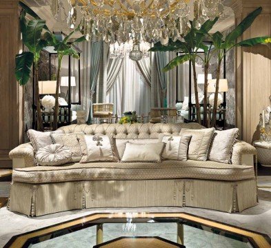 This picture shows a luxurious master bedroom designed with white and gold accents. The room features a large four-poster bed, two side tables with matching lamps, a comfortable sitting area, a patterned armchair, and a vanity area in the corner. There is also a chandelier hanging from the ceiling and a large window overlooking a beautiful garden.
