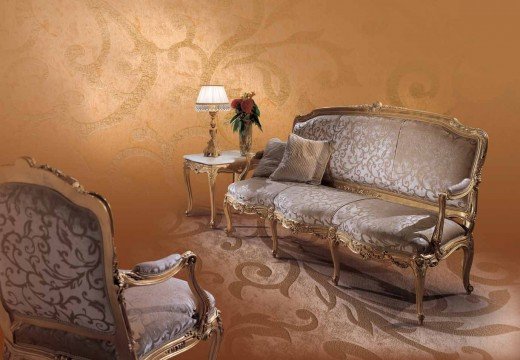 This picture shows a grand and luxurious living room. The walls are painted with a light grey colour and decorated with intricate golden designs. In the center of the room is a velvet tufted sofa in a creamy colour set against a patterned area rug. Behind the sofa is an ornate screen placed against a mirror with gold accents. There is a large crystal chandelier hanging from the ceiling, illuminating the room with a beautiful warm glow. On either side of the sofa are elegant chairs and small round side tables with detailed engravings. In addition, there are tall classical columns and statues