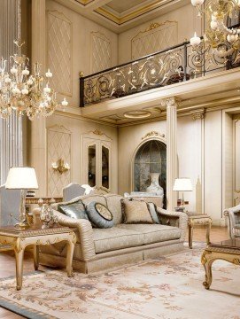 This picture shows a luxurious living room with a large cream-colored couch positioned in the center of the space. An intricate and ornate marble fireplace is against the wall behind it, and there are two large windows that offer plenty of natural light. The walls are white with gold accents, and a large golden chandelier hangs from the ceiling. There is an elegant round coffee table with a matching accent chair in front of the couch, and on the other side of the room there's a large wooden bookcase overflowing with books.