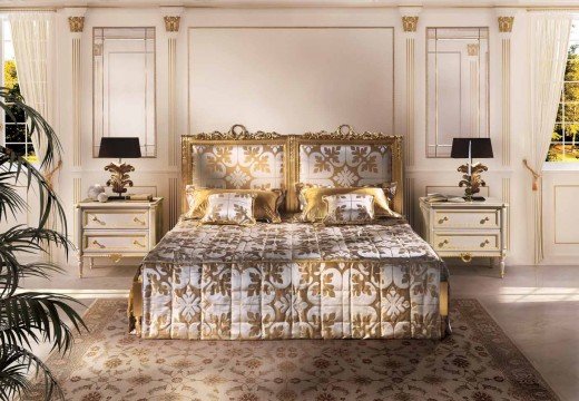 This picture shows a spacious bedroom with modern design elements. The walls are painted in a bright, light cream hue and are decorated with intricate moldings. The bed is large and upholstered in a pale beige color with tufted detailing along the headboard and footboard. The room also features a white side table with a lamp, a mirrored dresser, and a comfortable armchair. The decor includes several framed artwork pieces as well as an abstract metal sculpture.