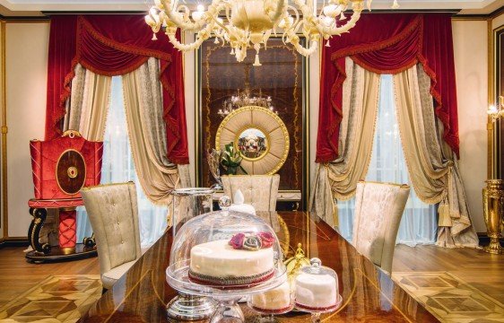 The picture shows an extravagant and luxurious interior design with a large staircase. The entire area is filled with white marble floors and walls. At the center of the room is a two-story room with a large, golden chandelier hanging from the ceiling. There are ornate gold railings and banisters that lead to the second floor, and at the top of the stairs there is a black and gold throne. On either side of the stairs, there are two beautiful statues of women dressed in white and holding up large lamps.