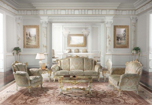 The picture shows a spacious, elegant living room space with modern and luxurious features. The room features a large cream and gray area rug, two cream colored couches, a matching armchair, and a beautifully decorated wooden coffee table. A round gold chandelier hangs from the ceiling above the furniture, and several pieces of art are hung on the walls. The overall effect is one of luxury and sophistication.