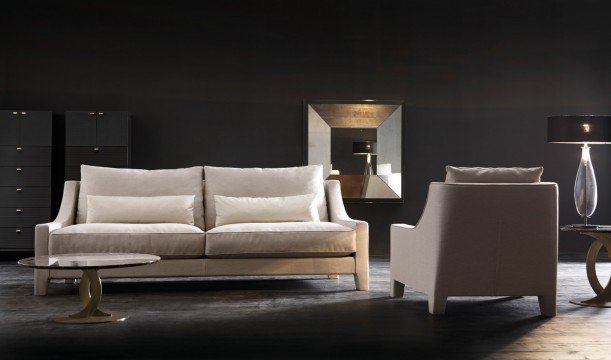 This picture is of an interior design project by Antonovich Design. It shows a modern, contemporary living room with a cream-colored sectional sofa. The walls are painted in a light beige color, and a patterned rug sits atop the tiled flooring. There is a glass coffee table in the center of the room, surrounded by four contemporary armchairs upholstered in a neutral-colored fabric. A large rectangular mirror hangs on one wall, and an abstract painting hangs above the sofa. A few plants are also seen throughout the space, adding a touch of greenery