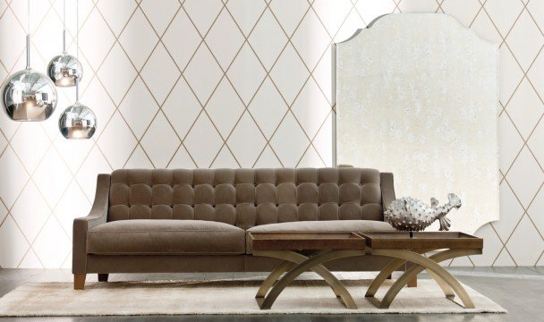 This picture is showing a luxurious open-plan living area with marble flooring and classic furnishings. It features a large white L-shaped sofa upholstered with comfortable cream cushions and decorative gold pillows, a glass coffee table, an elegant black and white patterned armchair, and two statement armchairs upholstered in velvet. The fireplace is situated on one wall and is framed by two stunning marble statues, while the walls are decorated with golden accents and pieces of artwork. There is also a chandelier above the room adding a touch of glamour.