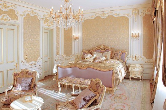 The picture appears to show a modern, luxury bedroom designed in shades of gold and white. It features a large king-sized bed with white bedding and a gold-colored headboard, a nightstand on either side of the bed, and a decorative rug beneath it. The walls of the room are lined with gold-trimmed white molding, and a large crystal chandelier hangs from the ceiling above the bed.