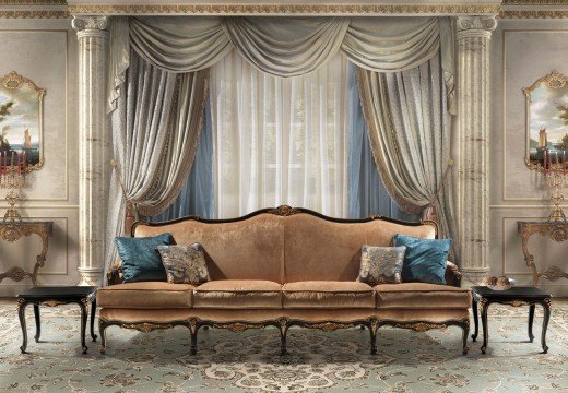 This picture shows an elegant and luxurious interior for a living room. The walls are painted in white and gold, and the floor is covered with a brown carpet with golden designs. The furniture is mostly white, with a large sofa upholstered with golden fabric, as well as two armchairs with pink and gold upholstery. On the opposite wall hangs a large golden mirror, which is framed with intricately carved wooden adornments. On the left side sits a tall white vase filled with fresh white and pastel-colored flowers. The ceiling is adorned with a crystal ch