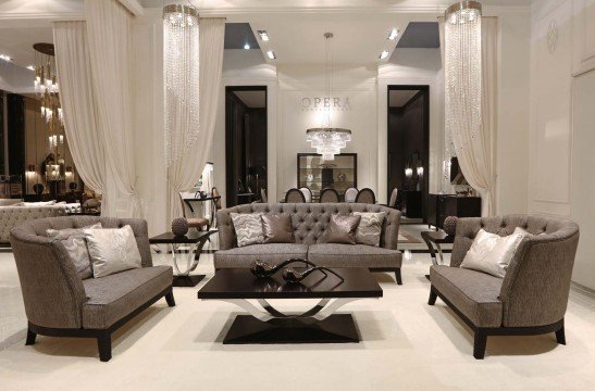 This picture shows an interior design scheme with contemporary furniture pieces in a spacious, predominantly white room. The walls and ceiling are painted a bright white while the hardwood floor has a light wood stain. A slate-gray velvet sofa is placed in the center of the room, with two matching armchairs and a luxurious ottoman arranged around it. On one side is a white marble fireplace, and on the other side is a large flat screen television mounted to the wall. The seating area is grounded by a diamond rug, and a tall wooden shelf filled with books gives the room a cozy feel
