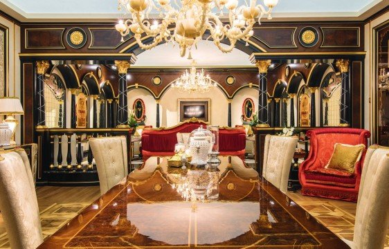 This picture shows a luxurious living space, with a crystal chandelier hanging from the ceiling and ornate furniture and fixtures filling the room. The wallpaper has a cream color with a swirl pattern, and there are large windows that provide plenty of light and a view of the outside. A grand staircase leads up to the second floor, and a dark-colored grand piano is visible in the corner.