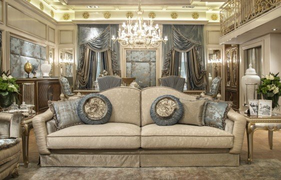 This picture is showing a grand, luxurious interior design in a wealthy home. The room features several large windows with ornate white curtains, an opulent chandelier hanging from the ceiling, and a plush cream-colored sofa with decorative pillows. There is also an elegant wooden coffee table in the center of the room. Various paintings are hanging on the wall and the floor is covered in a rich, patterned carpet.