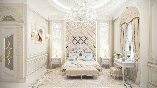 The picture shows a luxurious bedroom with an ornately-decorated ceiling and ivory-colored walls offset by a light grey floor. In front of the bed is a round wooden table with a delicate floral arrangement on top, and to the side of the bed is a large window with sheer curtains that is letting in natural light. There is also a sophisticated white armchair with a tufted back in the corner of the room, completing the look.