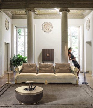 This picture is of an elegantly designed modern living room. The room contains a plush grey sofa, with white and pink patterned accent pillows, in the center of the room. There is a glass coffee table and a textured grey rug underneath. The walls are decorated with art pieces and a large circular mirror. On each side of the sofa there are grey armchairs with brass detailing, and behind the sofa is a wooden bookshelf full of books, plants, and other decorative items.