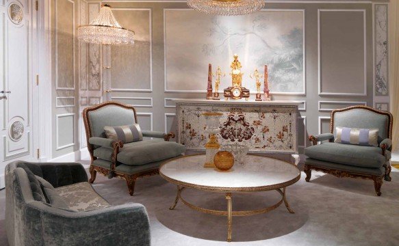 This picture shows an elegantly designed living room. It features a light beige couch with white and silver patterned throw pillows, as well as a glass coffee table that rests on a white and silver rug. The walls are painted a calming blue-grey color, and the floor is made of light colored wooden planks. There is also a large decorative mirror, some wall sconces, and an ornate chandelier hanging from the ceiling.