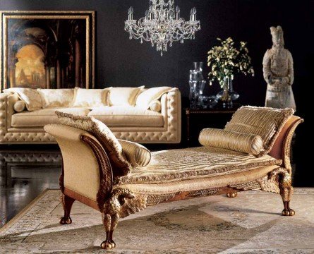 This picture shows an interior design featuring a luxurious living room. The main focus of the room is the cream-colored sofa and matching armchairs, arranged in an L-shape around a round glass coffee table. The walls and ceiling have been decorated with intricate gold accents, creating a warm and opulent atmosphere. A pendant light hangs from the center of the room, above the seating area. Several pieces of abstract artwork hang on the walls, adding a modern touch to the overall design. The floor is laid with a warm brown hardwood, and a large woolen rug provides additional warmth