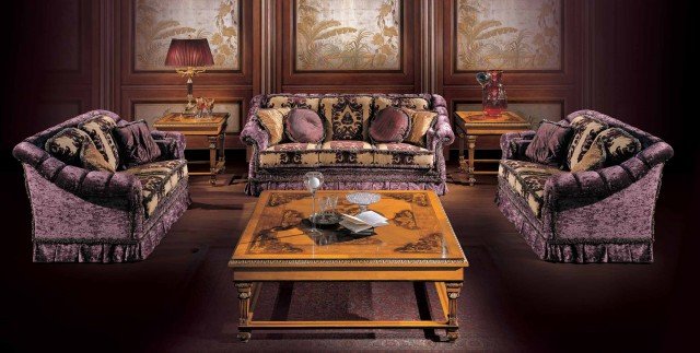 This picture shows a luxurious master bedroom with a wealthy decor. The space features a grand four-poster bed with an ornate gold frame and heavy cream curtains. On the right is a round end table with a lamp, and on the left is a chaise lounge. The walls are painted a deep red, while the floor is covered in a colorful Persian rug. The ceiling is decorated with a crystal chandelier and patterned wallpaper.