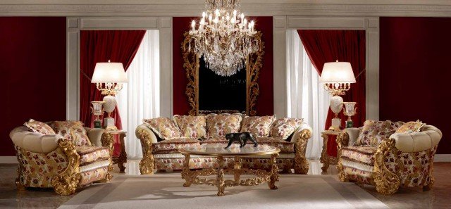 This picture shows a luxurious bedroom interior design. The room features a large bed with cream-colored bedding, a white tufted headboard, and gold accent pillows. There is a large window with sheer white curtains at the far end of the room, allowing natural light to flood in. The walls are painted a light gray with gold accent pieces on the wall. The room has built-in shelving for books and knick-knacks, as well as a couple of armchairs for relaxation. Finally, a large mirror hangs on the wall near the bed, reflecting the