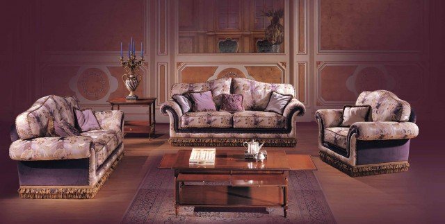This picture depicts a luxurious, modern living room. It features dark gray walls and an accent wall with ornate golden damask wallpaper. The furniture consists of a large, plush gray sofa, two side chairs in white upholstery, and a glass coffee table with a white base. There is a large framed mirror in the center of the wall and a chandelier with four cream-colored candles hanging from the ceiling. In addition, there are several decorative accents such as ornate gold wall sconces, framed artwork, and tall plants to complete the look.