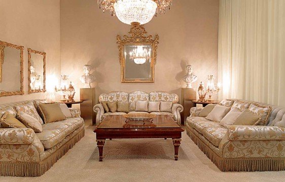 This picture shows an opulent and modern sitting room interior with grandiose chandeliers, large windows, high ceilings and a large fireplace with a cream-colored marble mantel. The walls are painted in a light grey and there are several stylish sofas and armchairs with luxurious gold-colored upholstery. On one wall, there is a modern painting depicting a stylized flower in shades of magenta and blue. The room has a large area rug with a white and gold pattern, and several green plants adding a touch of natural greenery to the space.