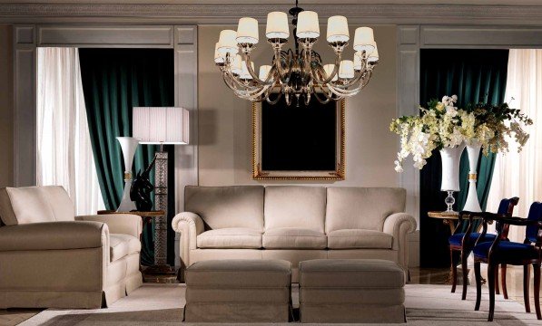 This picture shows a luxurious, modern interior design. It includes a white marble floor with a large crystal chandelier in the center of the room. There are several yellow velvet armchairs around the perimeter of the room, as well as a white sofa and glass coffee table. On the walls is a decorative wallpaper with blue and white geometric shapes. Large mirrors embedded in the wall also add to the contemporary appeal of the room.