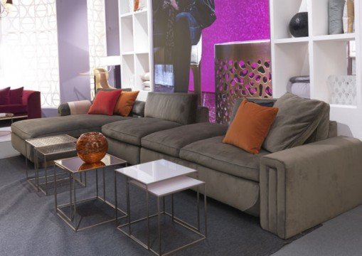 This picture shows a modern living room design with a black leather sectional sofa and a round glass coffee table. The walls are painted in a light gray color, and the floors are covered with large white and gray tiles. There is a white accent wall featuring a TV and shelf of decorations. A black and white chevron area rug provides contrast, while a tall potted plant adds a natural element to the room.