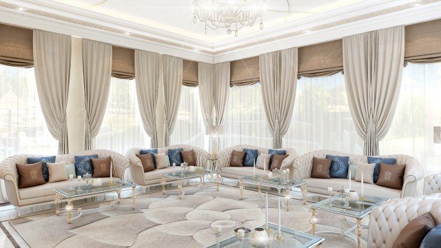 This picture shows a luxury living room with a beautiful crystal chandelier above the space. The room is filled with sophisticated and elegant furniture, such as an oversized white loveseat, navy blue armchair, white fur rug, and several gold-accented tables. On top of the tables are exquisite decorations, including a porcelain vase and a golden framed mirror. The walls are decorated with a golden and white patterned wallpaper and there are minimal plants placed around the room for decoration.