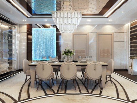 The picture shows an elegant dining room with a blue, velvet tufted banquette sofa and chairs surrounding a round glass top dining table, complete with a crystal chandelier. The walls are finished with white paneling and a large mirror is hung on one wall. The floor is covered with a thick, patterned area rug in shades of brown and gold.