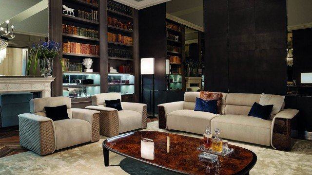 This picture shows the interior of a modern and luxurious living room. The walls are decorated with intricate wooden carved panels and beige-colored wallpaper. There are several lush armchairs with gold accents, as well as an ornate dark wood credenza. The centerpiece is a large white marble coffee table surrounded by an ornate sofa and two white lounge chairs. The room is illuminated by several gold and black chandeliers, while sunlight streams in through the large windows.