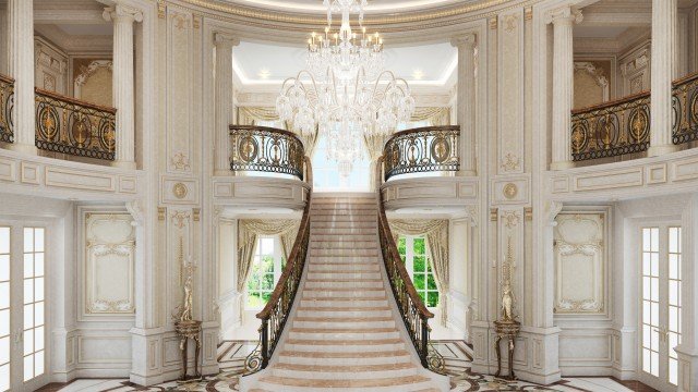 This picture shows a modern luxury stairway with white marble and brass accents. The stairway is illuminated by two large, sophisticated chandeliers, with glass panels and gold detailing giving the stairway a sophisticated look. There are two ornate handrails running along both sides of the stairway, and the steps are made of white marble and multiple colors of hardwood flooring. The walls are painted in a neutral tan color.