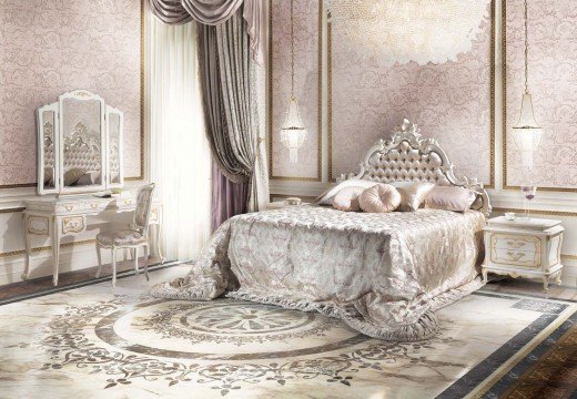 This picture shows a luxurious interior design of a bedroom. The walls are decorated with whitewashed wallpaper, and the room has a large four-poster bed with maroon and gold bed coverings. On the left side of the bed is an ornate wooden dresser with gold accents, and on the right is a white armchair. Above the bed hangs an elaborate chandelier, and in front of the bed is a wooden bench. The floors are covered in a beige and cream patterned carpet.