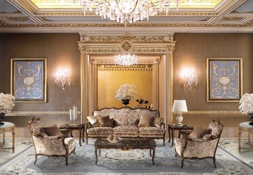 This picture shows a grand, modern living room with a classic, artistic flare. The room is decorated in beige walls and marble flooring, with several pieces of ornate white and gold furniture scattered throughout. In the center of the room, there is a large black marble table with a luxurious white chandelier hanging above it. A black and white art piece is hung on the wall to the right, and a cream-colored sofa sits in the middle of the room. There are two other doorways leading to other rooms, and a few potted plants scattered here and there to