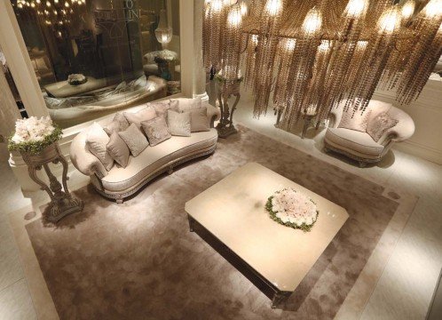 This picture shows an opulent, modern living room. The walls are painted pale grey and feature a few plain white floating shelves. In the center of the room is a large, contemporary-style white sofa with golden patterns. There is an oval light brown coffee table in front of the sofa, as well as two armchairs in a matching style. The room also includes a large, ornate, golden chandelier and two medium-sized crystal vases on the floor. Near the sofa, there is a white shelf, which is lined with books and other small items.