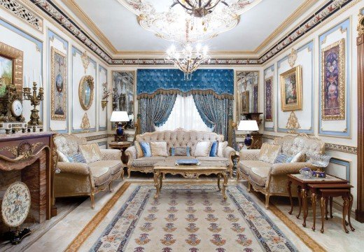 This picture shows a luxurious modern living room with beautiful gold details and rich, warm colors. The room features light grey walls, a white sofa, an ornate gold rug, an antique-style wooden coffee table, and two exquisite arm chairs upholstered in a deep purple fabric. The walls are adorned with large abstract artwork, and the space is finished off with two tall golden lamps.