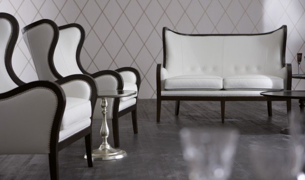 This picture shows an exquisitely designed living room. The room features a luxurious white leather sofa, accompanied by a sleek and modern white armchair. There is an intricate dark-wood coffee table with marble accents in the center of the room. The walls of the room are painted a soft gray color, with white and gold wall decor adding a shimmering quality to the interior design. A crystal chandelier hangs from the ceiling, illuminating the room and creating a warm and inviting atmosphere.