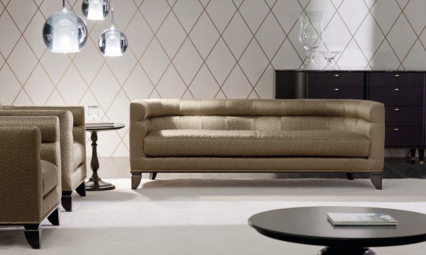 This picture shows a luxurious interior design featuring a modern living room with dark wood flooring and light beige walls. The room has a large flat-screen television mounted on the wall, several comfortable sofas and armchairs upholstered in light beige fabric and throw pillows in pastel colors, as well as accent pieces such as a glass coffee table and an area rug. The room also features a number of windows that are decorated with heavy drapery in a matching beige hue.