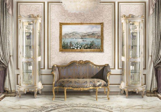 This picture is of a luxurious living room with cream-colored walls, marble flooring, and two plush white sofas. The room is furnished with a large glass coffee table, two armchairs, a rug, and several decorative plants. There are also two side tables with lamps and framed art on the wall.