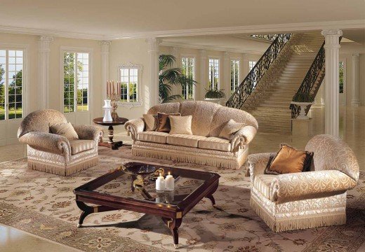 This picture shows a luxurious living room decorated with modern furniture and decorations. The main feature of the room is a large, ornate white sofa with pillows, set in the center of the room. The walls are painted a light color and have large windows that allow natural light to flood the space. The floor is carpeted in a white pattern, and the walls have some wallpaper and art hanging on them. A sleek, glass table with gold accents is at the center of the room, and there are several other pieces of furniture scattered around the room.