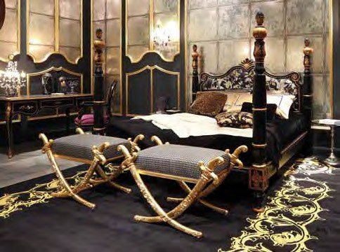 The picture shows an ornate, modern-luxury bedroom. The room has a taupe-colored wall with a white crown molding and a sunburst mirror. Against the wall is a curved headboard on a white bed adorned with several throw pillows. The bed is flanked by two glass-topped nightstands with white drawers. A silhouette wall-art piece is hung above the bed and a cream area rug covers the floor.