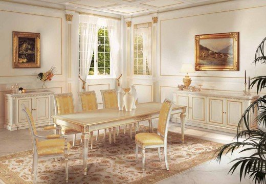 This picture shows a luxurious living room with white and gold accents. The room includes a white leather couch, two gold arm chairs, a white coffee table with a glass top, and a large cream area rug. The walls are decorated with white panels and gold trim, and a large gold frame mirror hangs on one wall. Recessed lighting is installed in the ceiling and floor-to-ceiling windows allow for plenty of natural light.