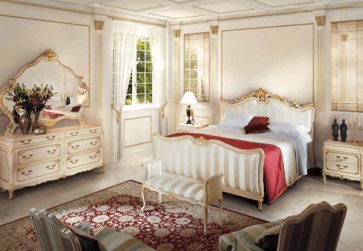 This picture shows a large, luxurious bedroom with a coffered ceiling and walls decorated with intricate molding and gold accents. The bed is a four-poster frame with white bedding and dark wood detailing. On the left side of the room, there is a tall, cream and gold armoire, and beside the bed is a nightstand with built-in drawers. The floor is covered by a patterned area rug in shades of brown, green and yellow.