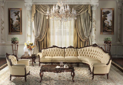 This picture shows a luxurious living room with modern interior design in shades of white, silver and light beige. The room is elegant and spacious with a glass-topped coffee table, two large sofas upholstered in white and pale beige fabric and a set of two white chairs. A grand chandelier hangs from the high ceiling, while a patterned area rug adds warmth to the hardwood flooring. High windows allow natural light to flood into the room. The walls are decorated with modern wall art.