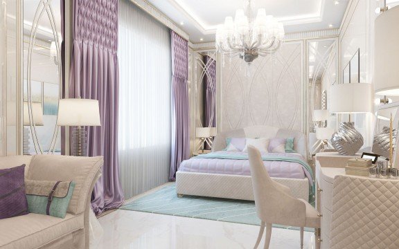 This picture shows a luxurious bedroom in a residence. The walls and ceilings feature ornate white and gold trim, with an elegant chandelier hanging from the center of the room. There is a large bed in the middle of the room, with a soft white and gold bedspread, and several throw pillows. To the side of the bed is a sitting area with two white chairs, a small table, and a golden mirrored end table. The floor is covered in a light beige carpeting. Hanging on the wall are several framed pieces of art.