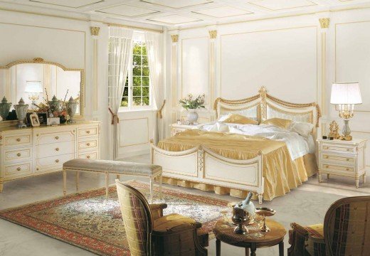 This picture shows an impressive and luxurious bedroom with a classic design. The walls are painted a light beige color, the ceiling is elaborately decorated with rosettes, and the furniture has deep blue accents. The bed is placed in the center of the room, surrounded by two nightstands and two armchairs. A large crystal chandelier hangs from the ceiling, which further showcases the elegant design.