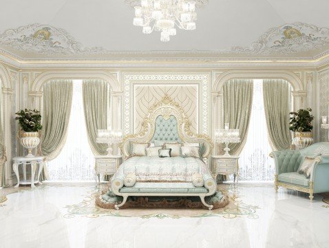 This picture shows a lavish interior design in a living room. It features a large, beige tufted leather sofa with gold accent pillows, a maroon ottoman, and a round glass-topped coffee table. The walls are painted a rich, eggshell color, while the ceiling boasts a regal, crystal chandelier. On one side of the room is a stately, wood mantel with an ornately framed mirror above it. On the other side is a bar area framed by tall, built-in dark wood cabinets. Various art pieces hang on