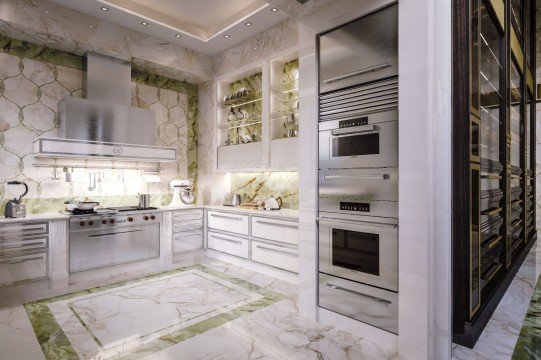 This picture shows a luxurious kitchen with a marble countertop and backsplash, two separate islands, and white cabinets and drawers. There is a built-in refrigerator and a large double oven. The kitchen also includes plenty of natural light, as well as two modern hanging lights. The entire space is finished with a white stone floor.