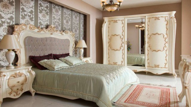 This picture shows a luxurious bedroom interior with a modern yet classic design. The light-colored walls and the dark wooden floors contrast nicely with one another, while the large window provides ample natural lighting. The furniture is a mix of classic and modern pieces, including a tufted luxurious headboard, a matching armchair, and a white nightstand. The intricate patterned wallpaper, along with the gold tones in the armchair and bedding, provide a touch of elegance and sophistication to the room.