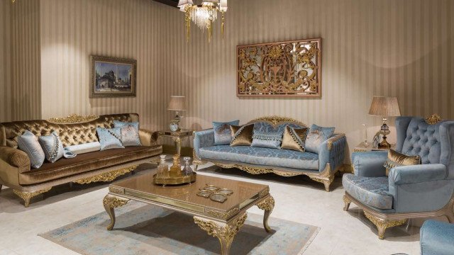 This picture shows an elegant and luxurious living room. The walls are painted in a soft gray hue with white trim, and the floors are covered in a cream-colored carpet. In the center of the room is an oval-shaped light fixture that hangs over a curved sofa. On either side of the seating area are two ornately detailed armchairs with gold accent pillows. The wall behind the sofa is adorned with two large mirrors, while the wall to the side of the doors is decorated with decorative wallpaper. An intricately detailed glass coffee table completes the look.