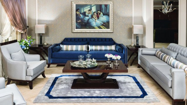 This picture shows a modern luxury living room designed by Antonovich Design. This room features elegant furniture, such as a sofa, coffee table and armchair, all in white leather. There is a beautiful crystal chandelier hanging from the ceiling, as well as a large flat-screen television mounted to the wall. The walls are painted in a light shade of grey and the flooring is made of dark marble.